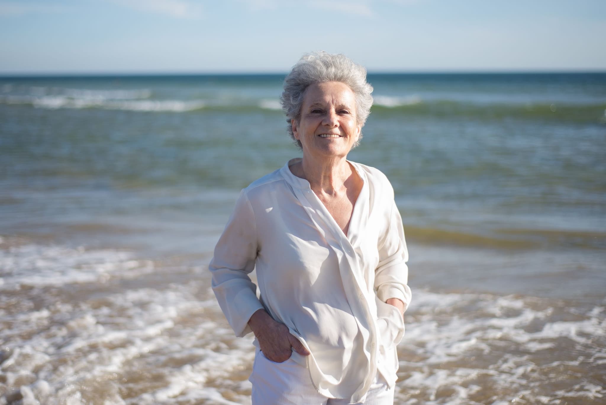 Nasmiješena starija žena u bijeloj košulji stoji na plaži, iza nje more i valovi